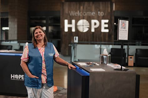 A West Des Moines volunteer stands under the New to Hope area wearing a blue vest smiling at the camera