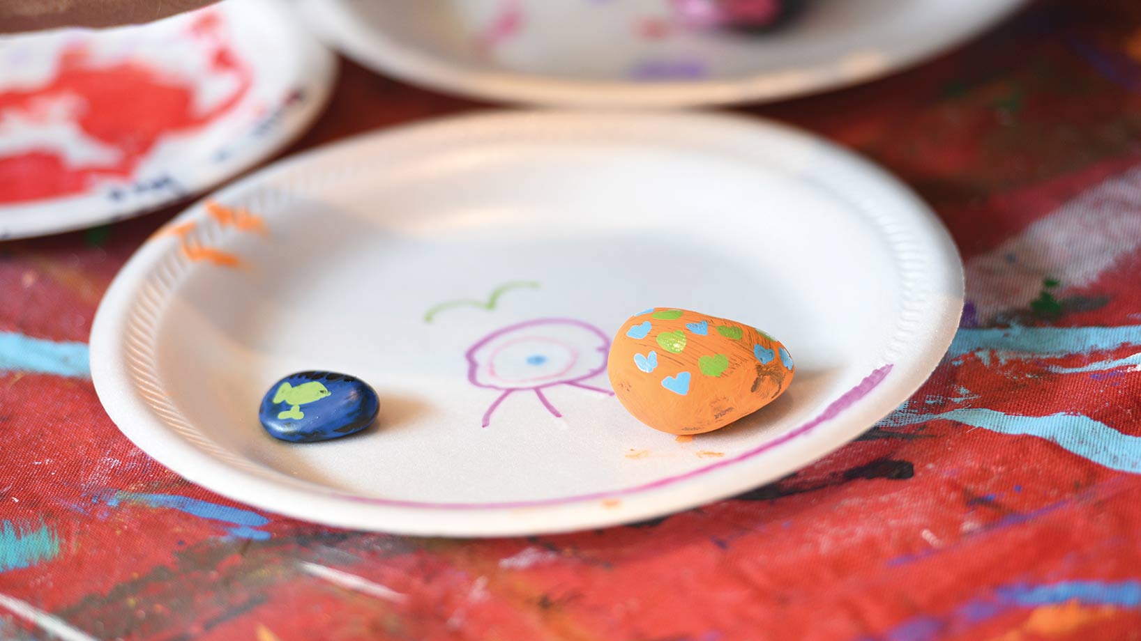 Two painted rocks on a plate with doodled drawings. Other plates seen in background, all on paint-covered tablecloth