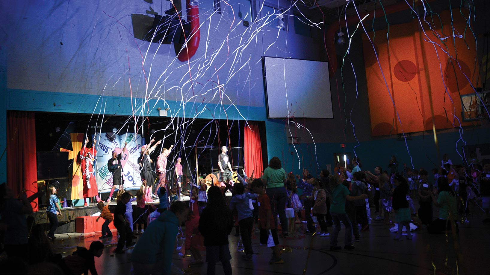 Kids dancing in gym with adults dancing on stage with streamers in the air