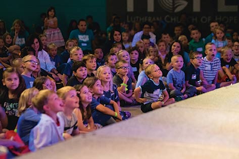 Young kids sitting excitedly watching something on stage in the gym