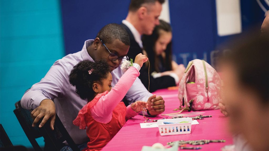 Dad and daughter work on making a bracelet together at the table.