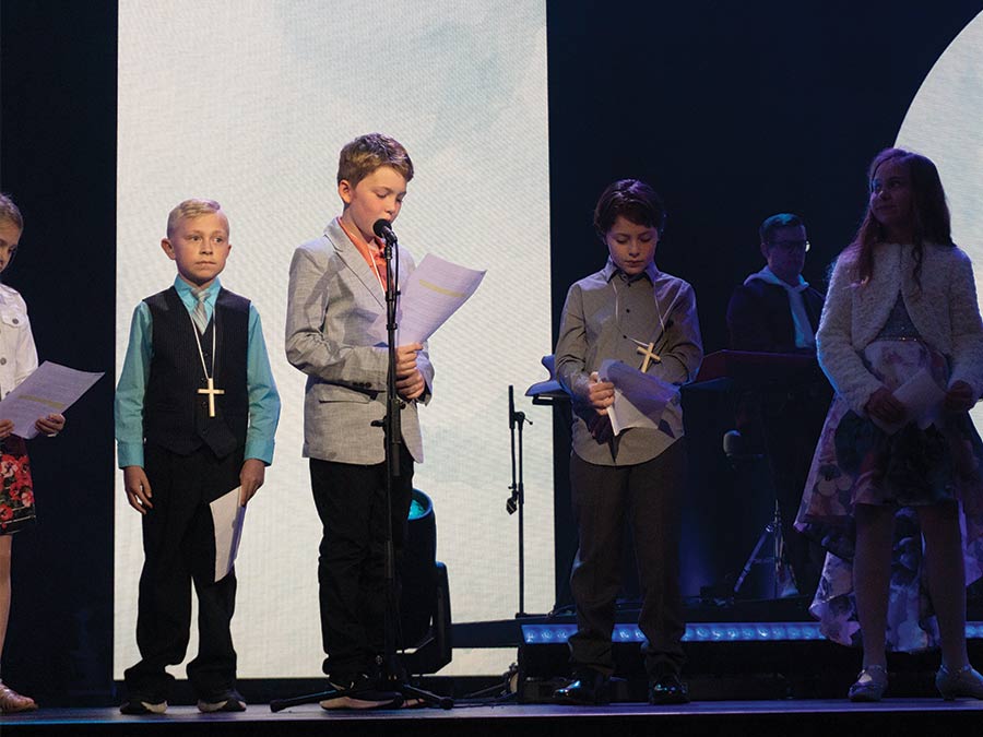 Kids standing on stage in nice outfits with papers in their hands and wearing large cross necklaces.