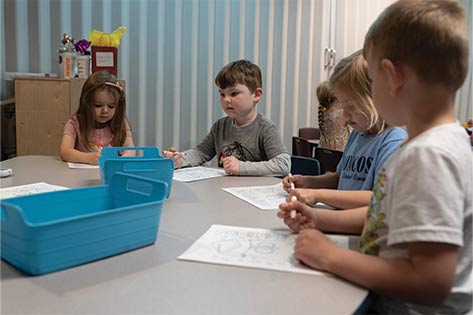 Group of students sit at table with coloring sheets and markers in hand