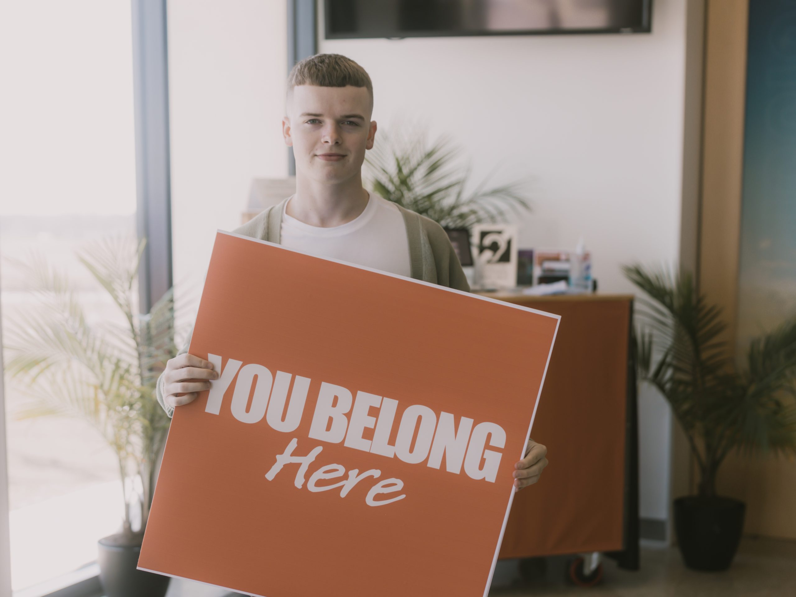 Leader holding an invitational orange sign in the Hope Grimes building that reads "You Belong Here"